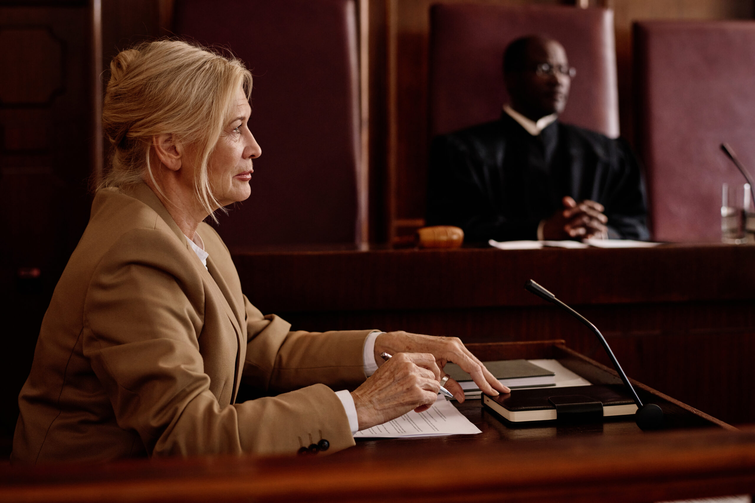 Side view of mature blond female witness or lawyer speaking in microphone while sitting against male judge